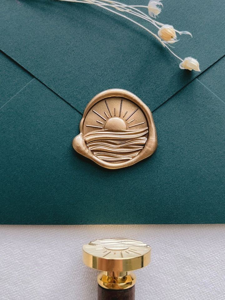 Gold 3D sunset over ocean waves wax seal on a dark green envelope next to a wax stamp head with dark wooden handle