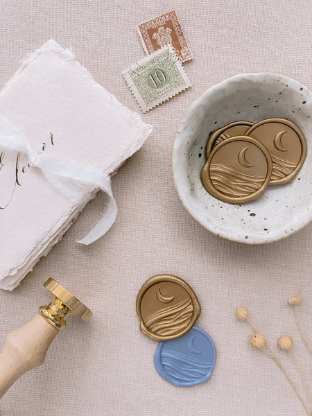 3D moonlight wax seals in gold and dusty blue styled with gold wax seals in small bowl, postage stamps, place cards tied with white silk white ribbon and wax seal stamp