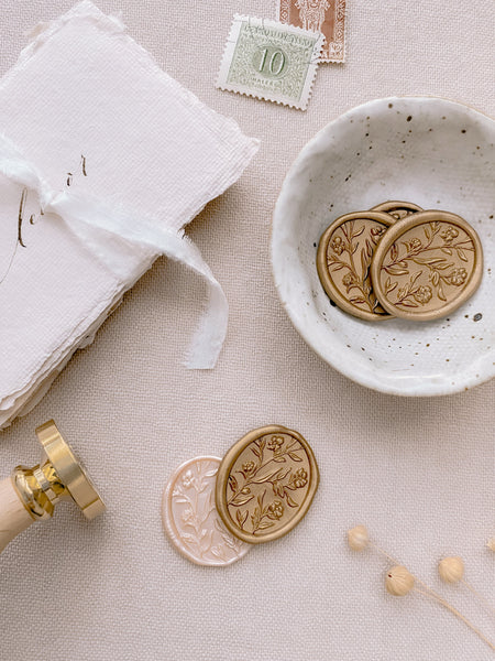 Oval floral wax seals with 3D engraving in light gold and pearl styled with gold wax seals in small bowl, place cards tied with white silk ribbon, wax seal stamp and postage stamps