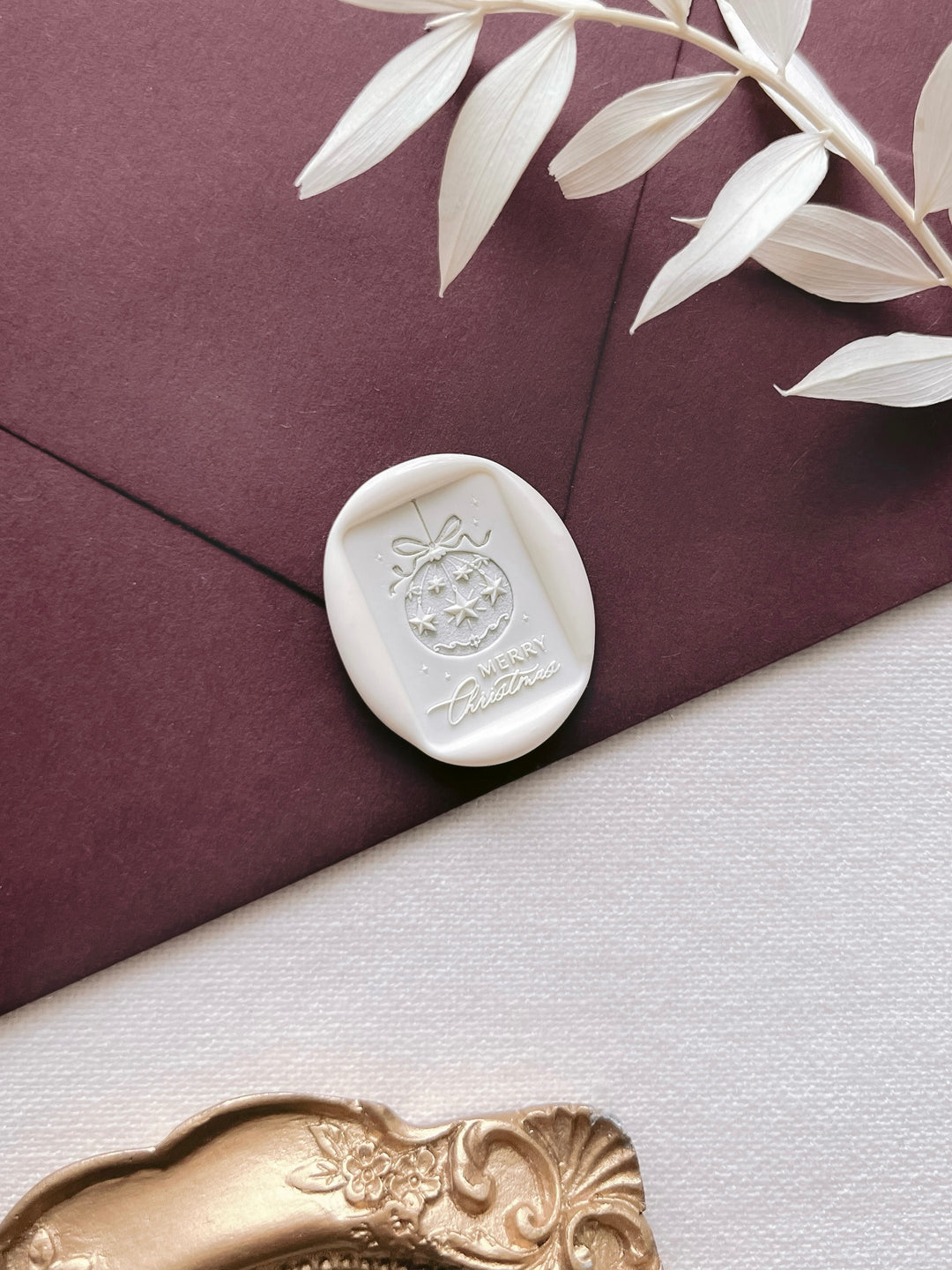 a white wax seal with a decorative design, placed on a dark surface next to a branch with white leaves.