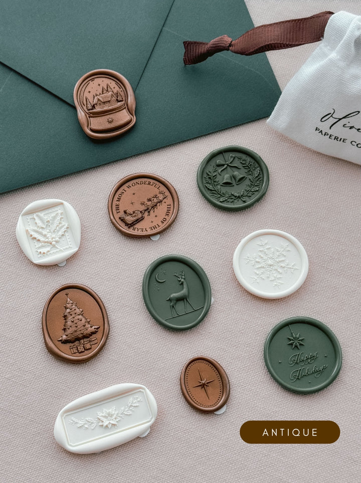 Assorted Christmas wax seal stickers in bronze, dark green and ivory — ten festive designs arranged beside a dark green envelope and a linen drawstring pouch.