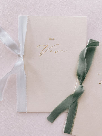 A set of two His and Her gold foil beige card stock vow books in calligraphy script with one tied in white colored silk ribbon and the other in olive green silk ribbon