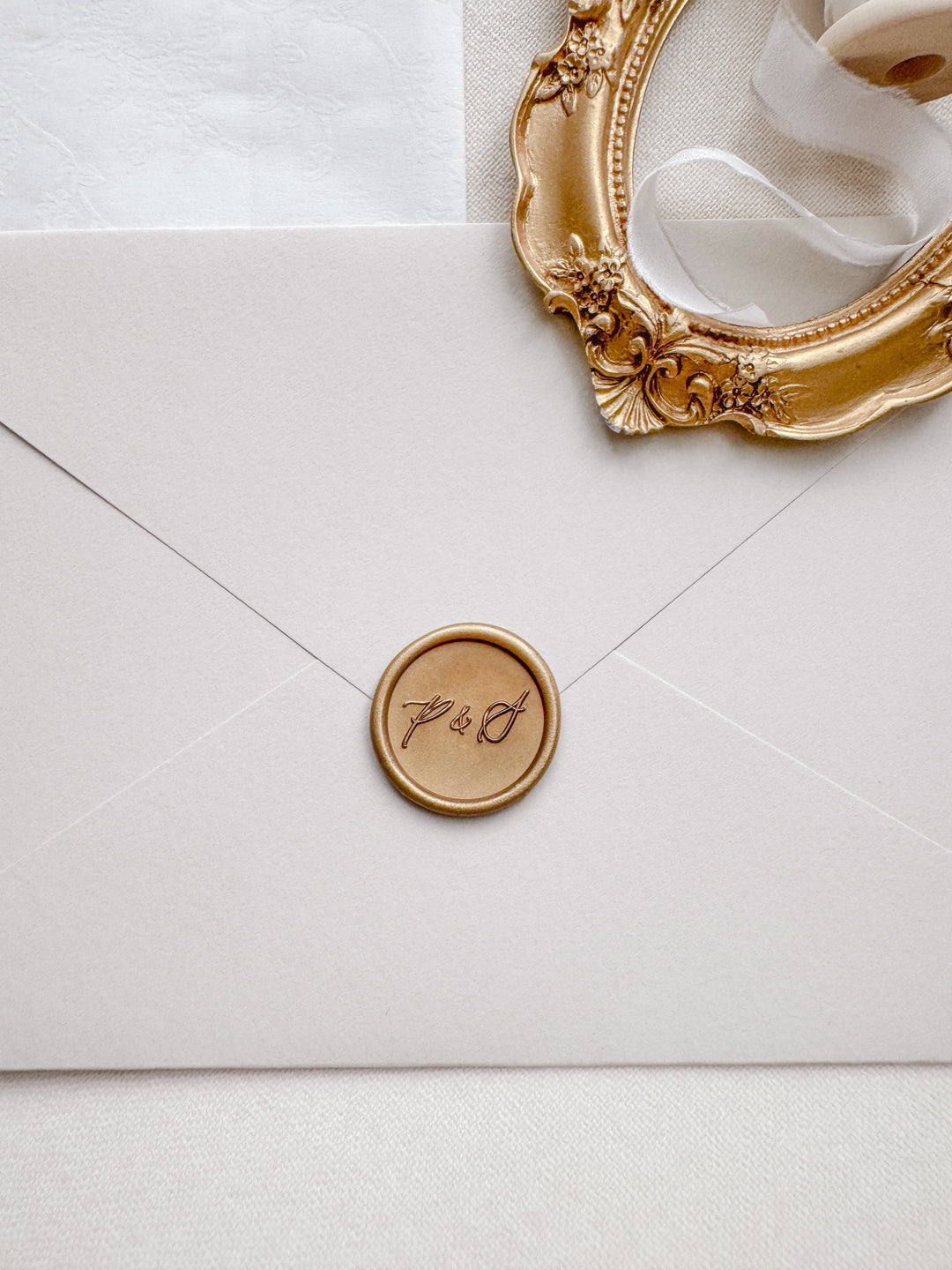 Calligraphy script monogram gold wax seal on a beige envelope styled with a gold vintage oval frame prop