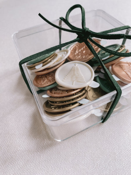 A box of assorted designs and colors in antique white, sage, light gold and gold Christmas wax seal stickers wrapped with Christmas dark green suede ribbon side view