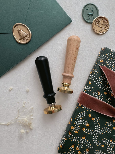 Christmas wax stamp kit stamp handle options in wooden color and black, next to Christmas card with Christmas tree wax seal and wrapped gift