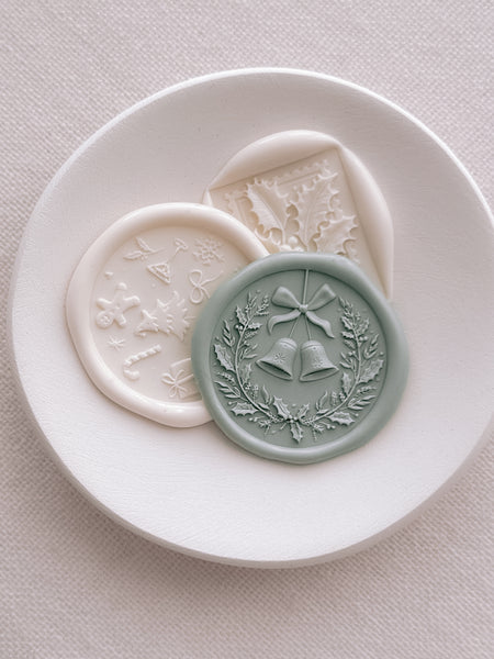 Light sage green Christmas wreath and jingle bells wax seal, off-white festive symbols wax seal and an off-white holly leaf wax seal on a small dish