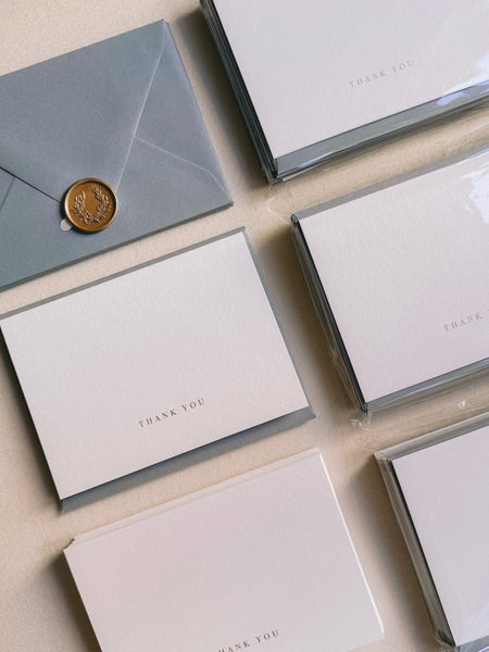 Stacks of gold foil white thank you cards with dusty blue envelopes and gold wax seal