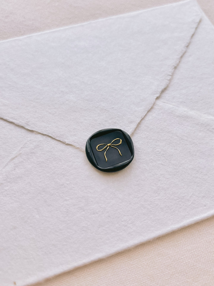 square shaped hand-drawn bow design wax seal in black with the design painted in gold 