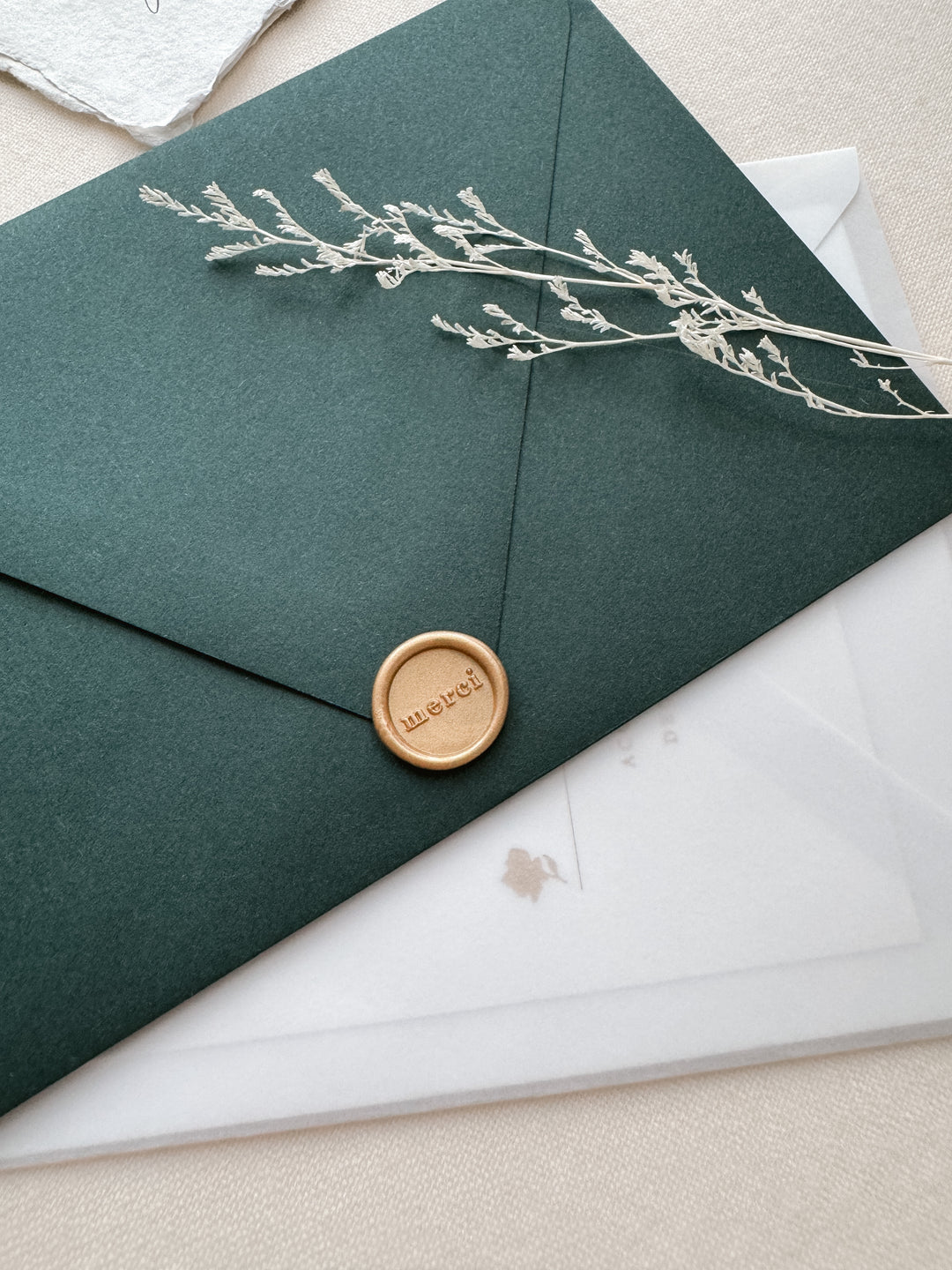 Light matte gold mini merci wax seal on a dark green envelope on a white wedding invitation vellum envelope