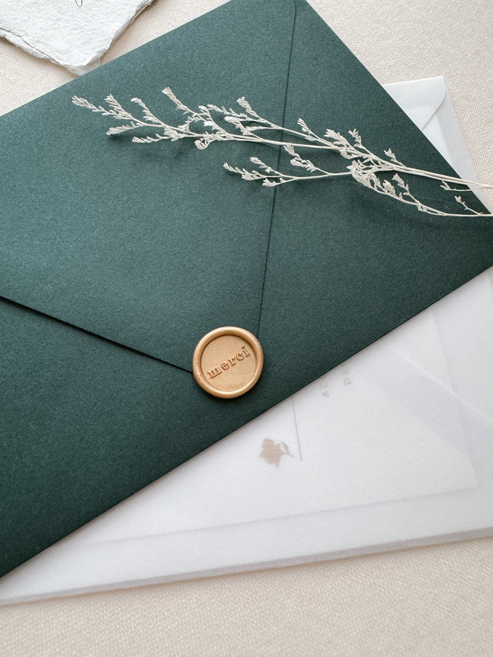 Light matte gold mini merci wax seal on a dark green envelope on a white wedding invitation vellum envelope