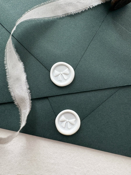 White mini ribbon bow wax seals on dark green envelopes styled with a strand of white silk ribbon