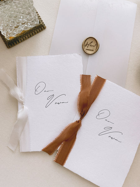 A set of two His and Her letterpress OUR vow books in calligraphy script with one tied in antique white colored silk ribbon and the other in cinnamon colored silk ribbon styled with vellum wrap and gold wax seal