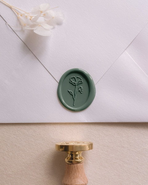 Oval floral wax seal in dark sage green on a white paper envelope styled with a wax seal stamp and dried flowers