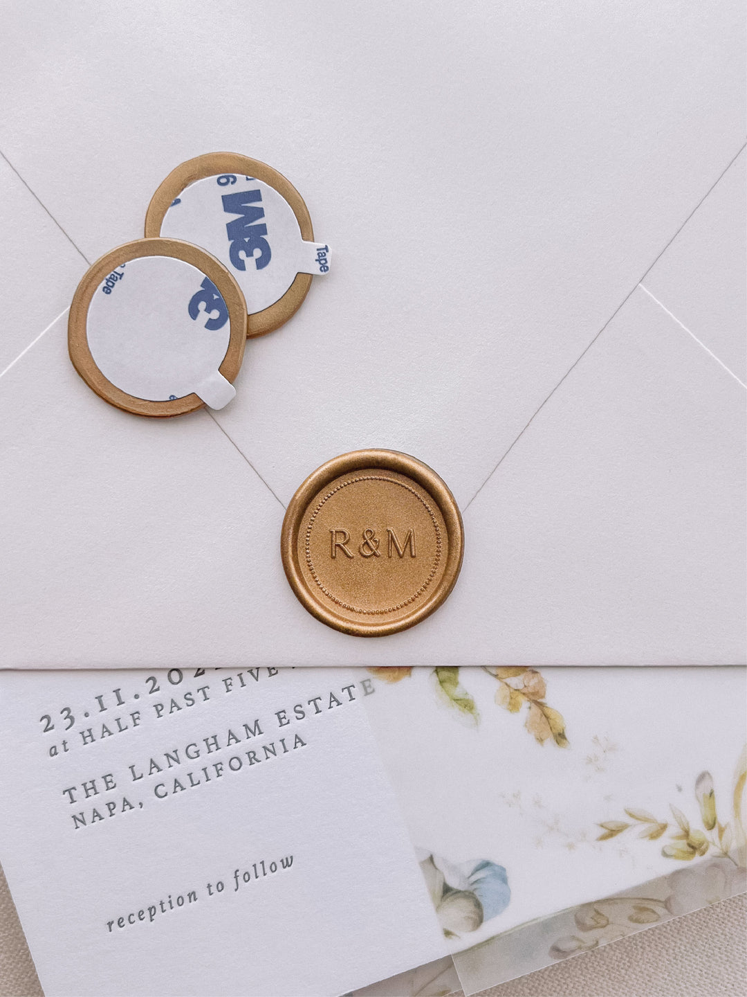 Gold custom monogram wax seal stickers with a dot border design, with self-adhesive backings on a wedding invitation card, beige envelope, and vellum jacket