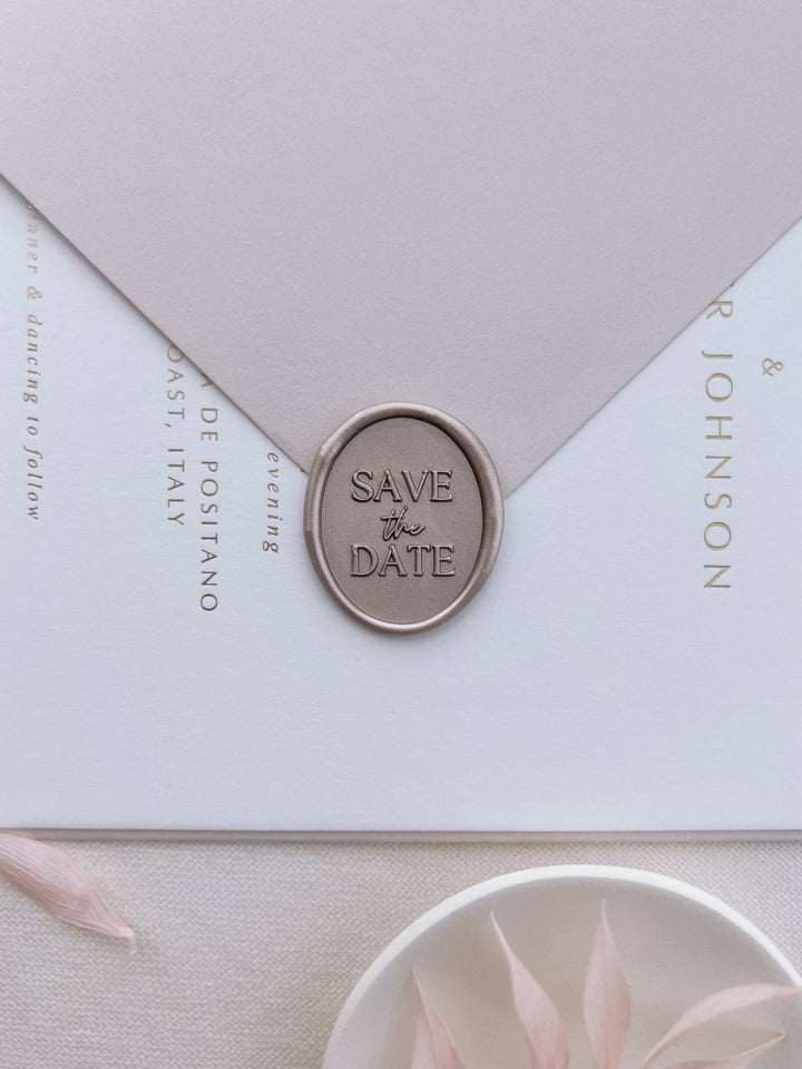 Light bronze save the date oval wax seal on a light beige envelope over a gold foil wedding invitation card 