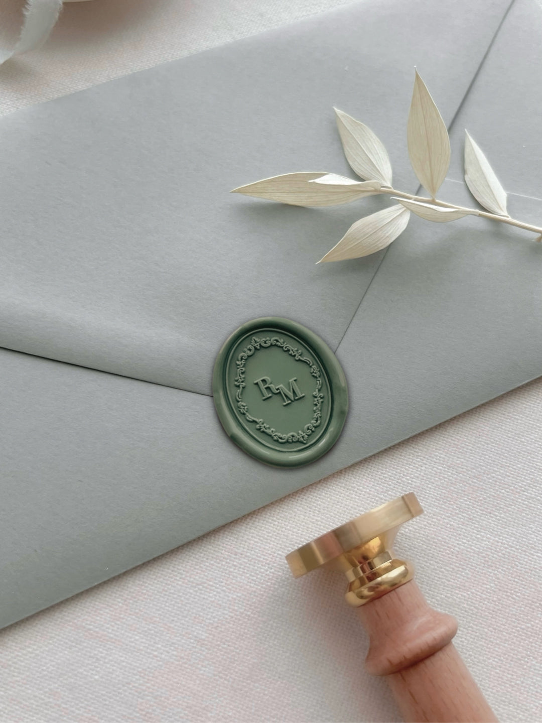 Green vintage Baroque crest oval wax seal with personalized initials on a light sage green envelope next to a wax stamp head with wooden handle