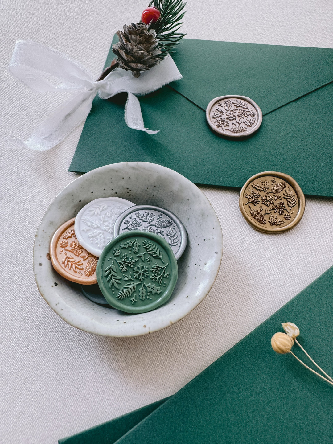 Festive wax seals in various colors featuring snowflake, botanical and floral elements, placed in a small ceramic dish and on a dark green envelope