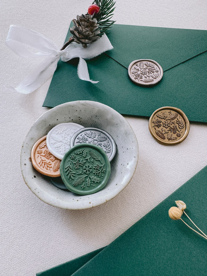 Festive wax seals in various colors featuring snowflake, botanical and floral elements, placed in a small ceramic dish and on a dark green envelope