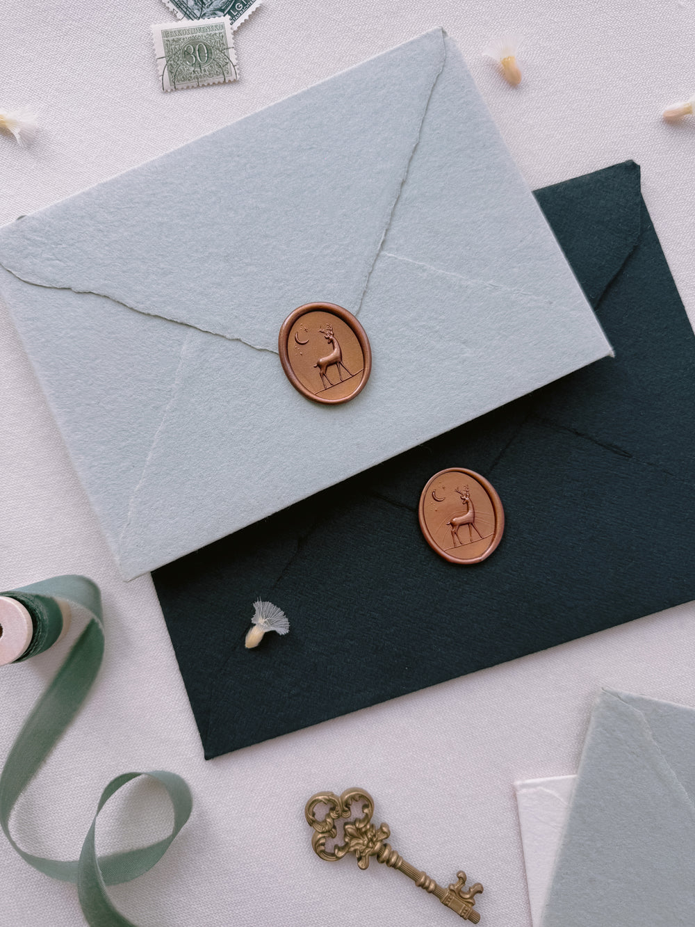 Dark metallic bronze 3D reindeer oval wax seals on a light blue and black deckled edge handmade paper envelopes on a light beige background