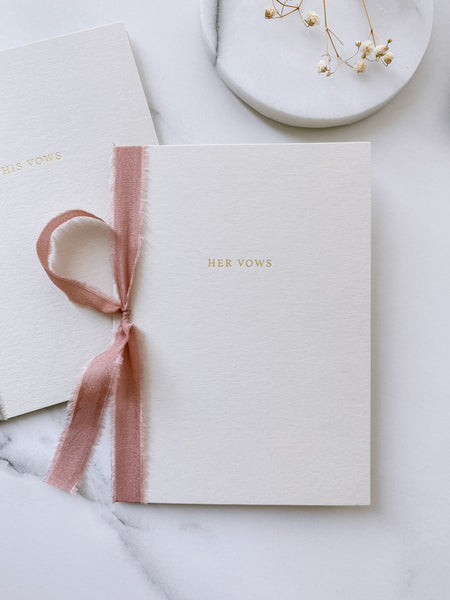 A set of two His and Her Vows beige card stock vow books in typeface font printed in gold foil tied in dusty rose colored silk ribbon