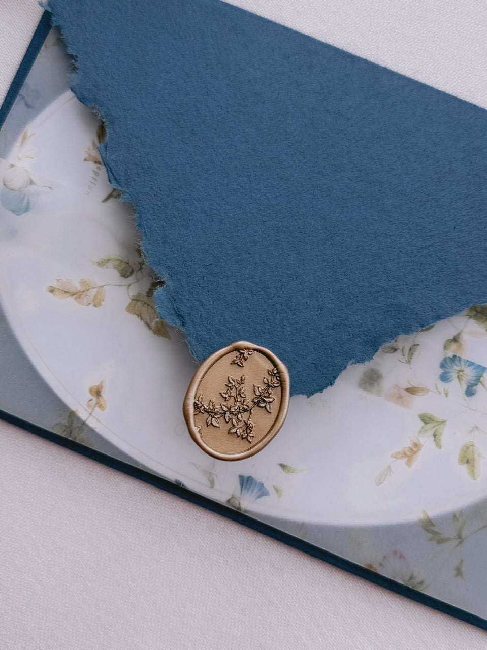 Gold botanical vine oval wax seal stamp on a deckle-edge blue envelope, over a floral print wedding vellum jacket