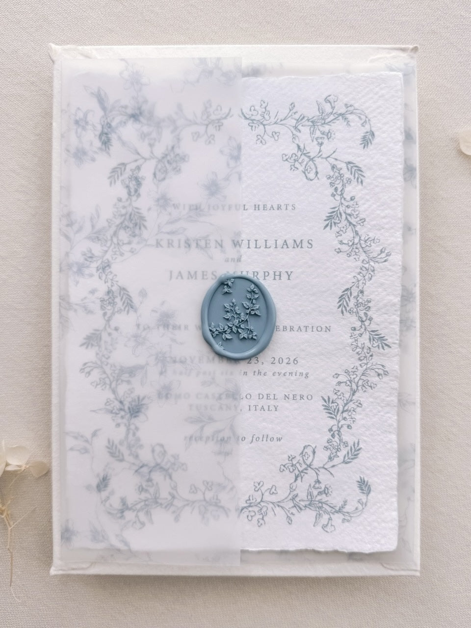 Dusty blue botanical vine wax seal on a vellum overlay, layered over a hand-illustrated floral wedding invitation card.