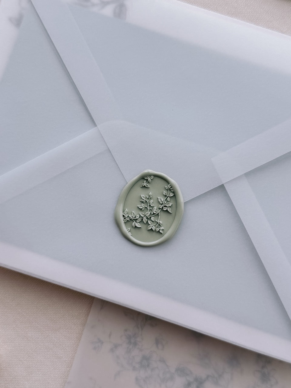 Sage green botanical vine oval wax seal on a vellum envelope set against a blue floral print vellum wrap