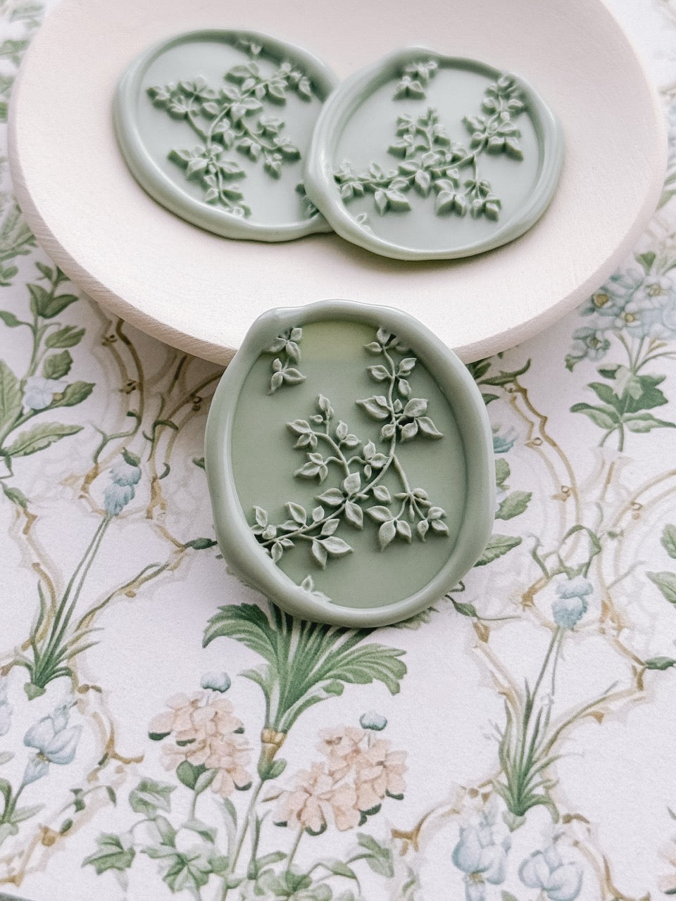 Sage green botanical vine wax seals displayed on a ceramic dish, set against a vintage floral backdrop.