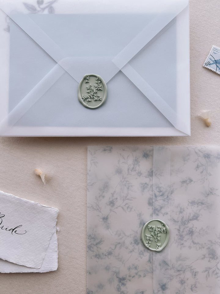 Sage green botanical vine oval wax seal on a vellum envelope set against a blue floral print vellum wrap