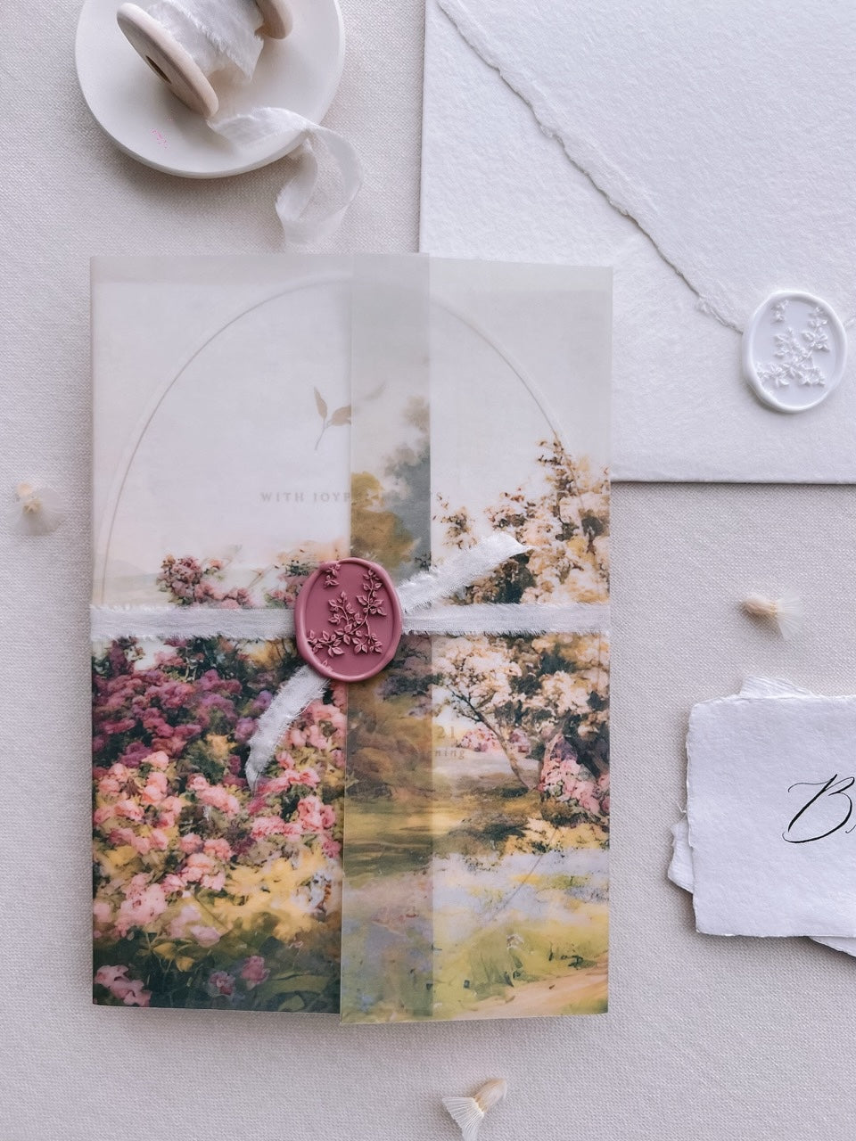 Mauve pink botanical vine wax seal securing a ribbon-tied watercolor garden print vellum overlay on a wedding invitation, with a white wax seal on the white envelope behind.
