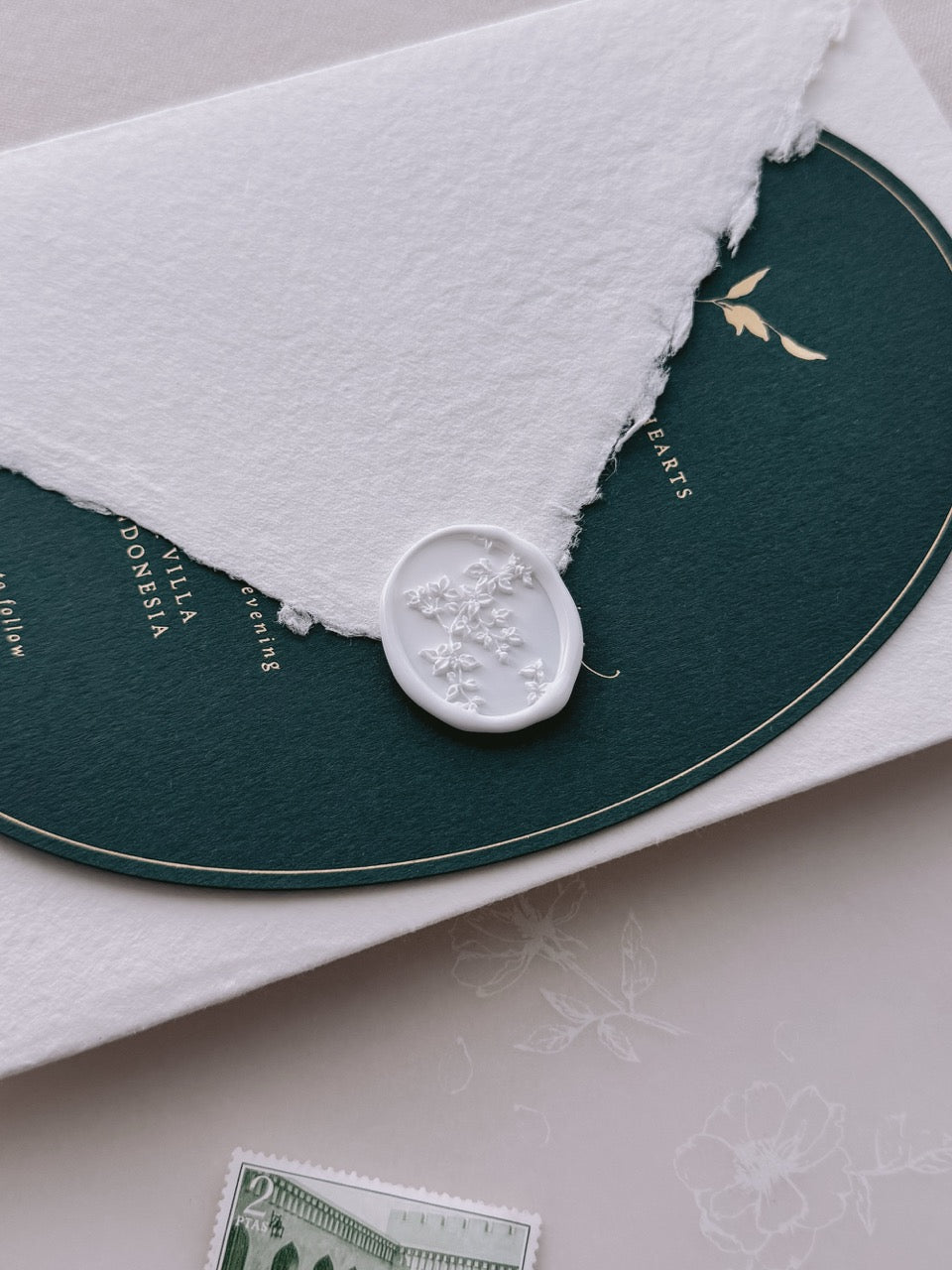 White botanical vine oval wax seal on a deep green wedding invitation card with gold foil detail, set against a deckle-edge envelope.