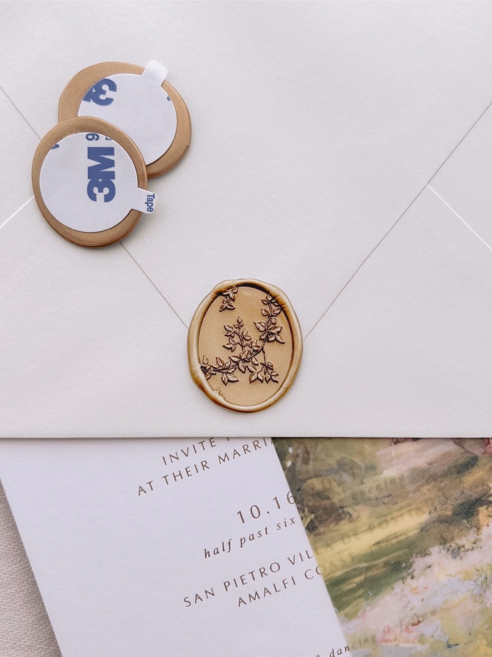 Gold pre-made botanical leaf oval wax seal stickers with self-adhesive backings on a beige envelope over a white wedding invitation card and a watercolor vellum jacket