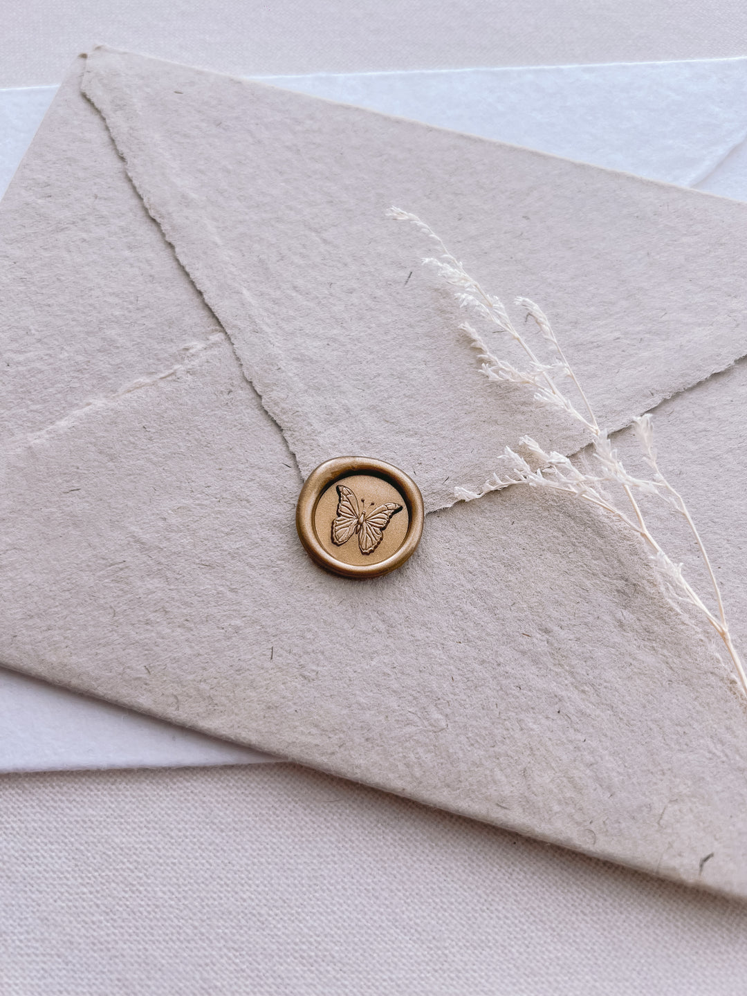 gold 3D butterfly mini wax seal on an oatmeal deckle-edge handmade paper wedding envelope with dried grass