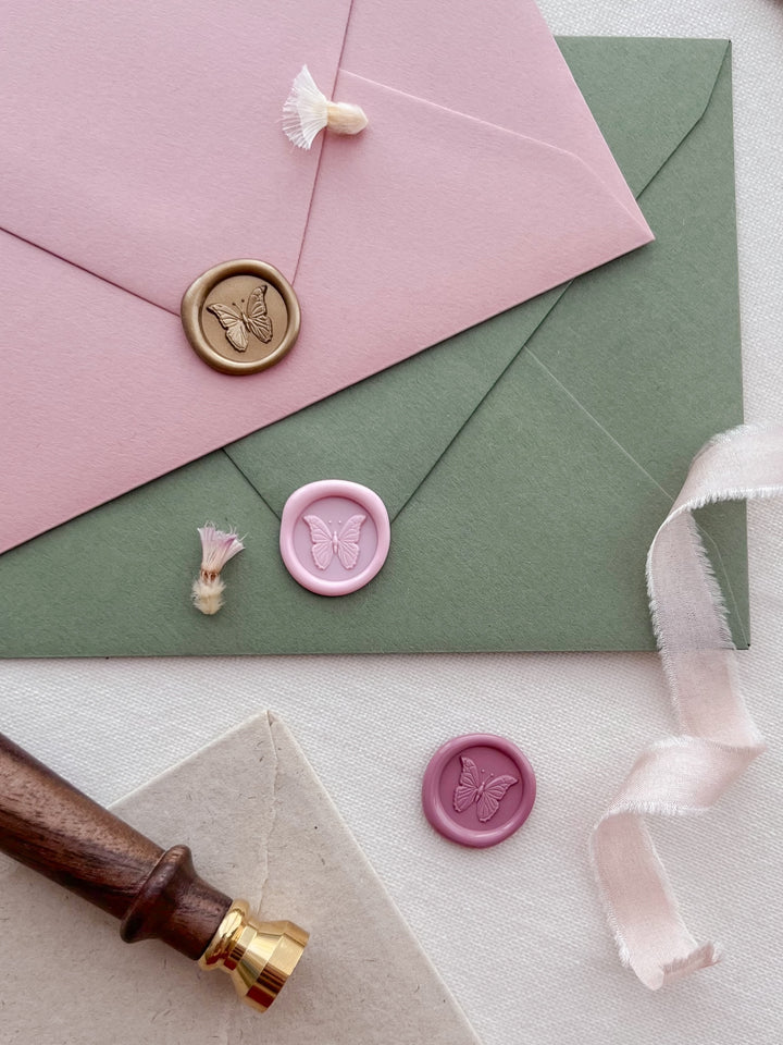 light gold, light pink, and rose pink 3D butterfly mini wax seals on a dusty rose and moss green wedding envelopes with white silk ribbon and a brass stamp handle