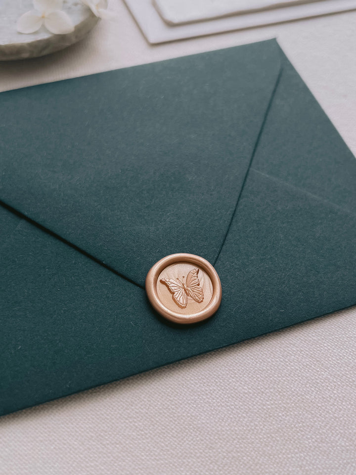 gold 3D butterfly mini wax seal on a dark green textured wedding envelope and white florals