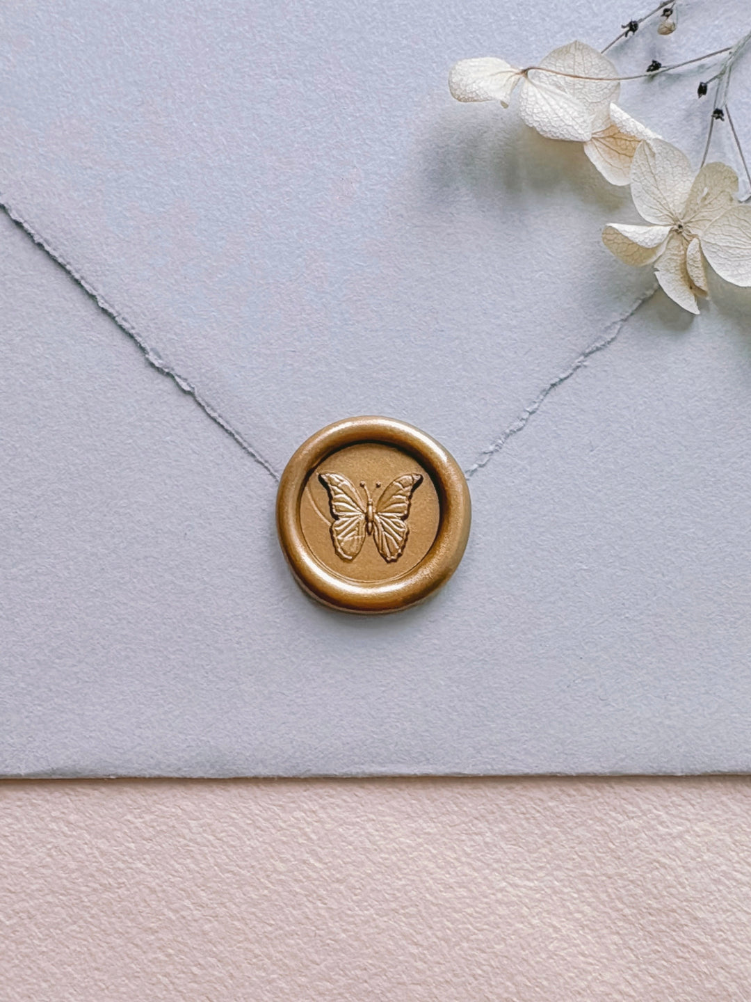 light gold 3D butterfly mini wax seal on a flap of a light blue deckle-edge wedding envelope with dried hydrangeas