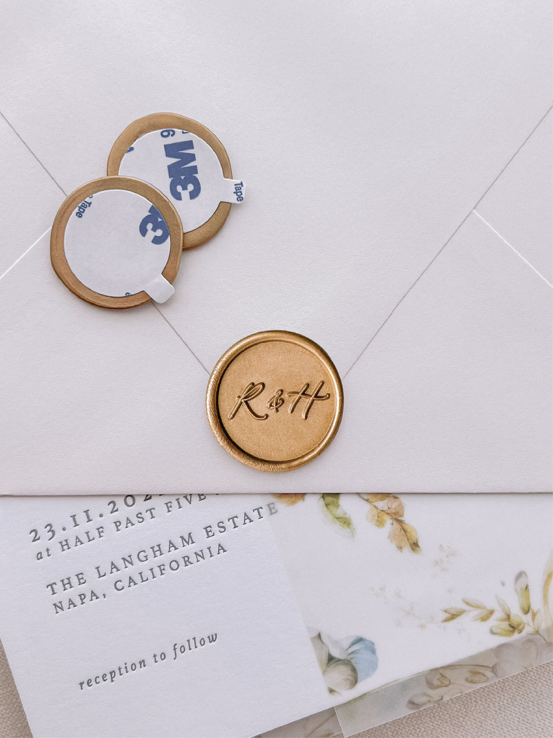 calligraphy monogram round with "&"
 gold wax seal stickers with self-adhesive backings on a wedding invitation, beige envelope, and floral vellum jacket