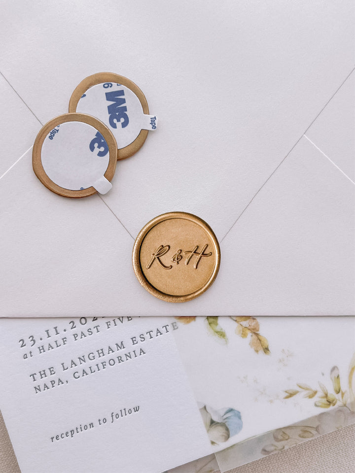 calligraphy monogram round with "&amp;"
 gold wax seal stickers with self-adhesive backings on a wedding invitation, beige envelope, and floral vellum jacket