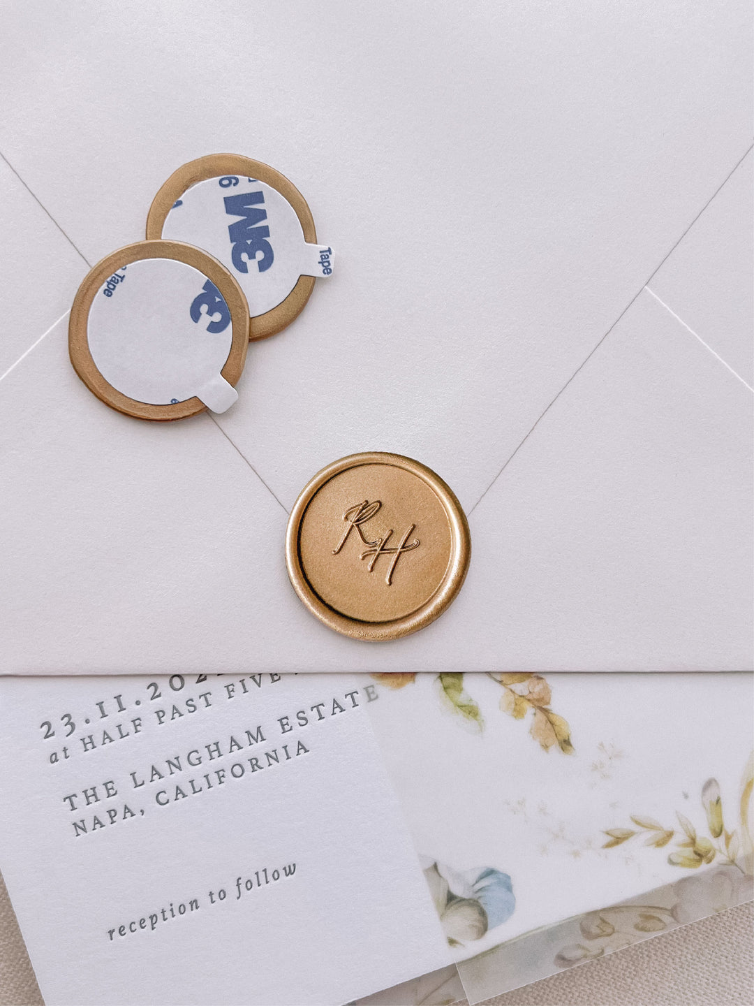 calligraphy monogram round gold wax seal stickers with self-adhesive backings on a wedding invitation, beige envelope, and floral vellum jacket