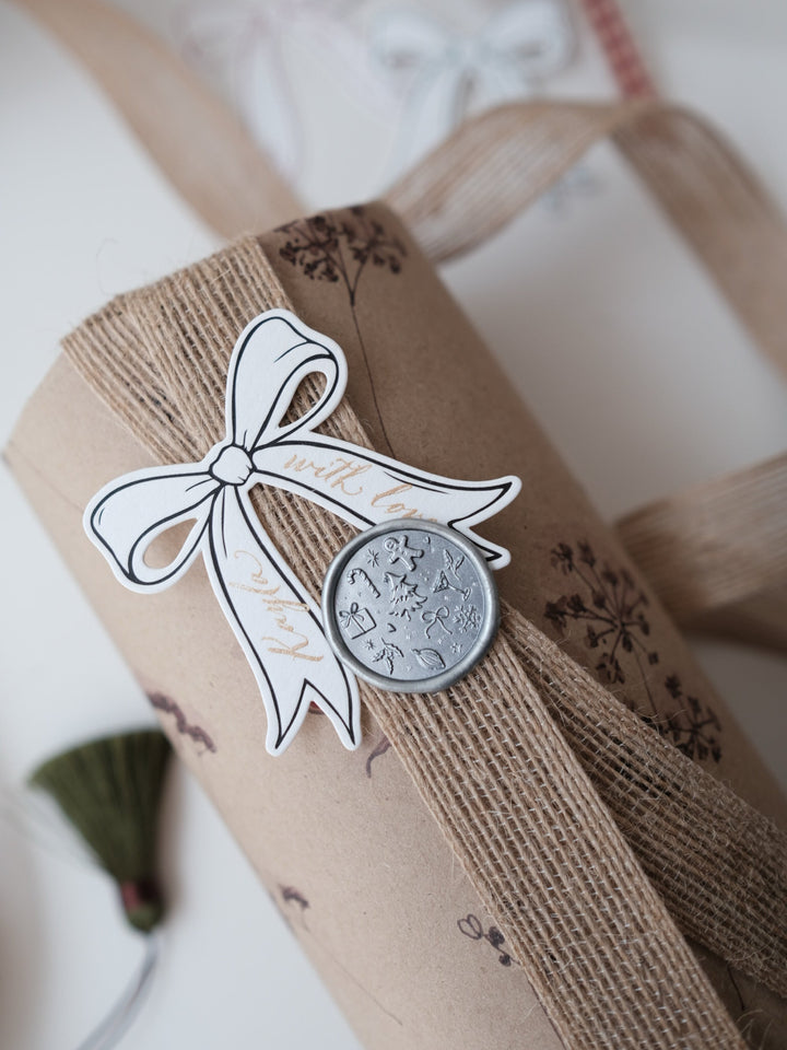 Silver holiday wax seal with festive icons and symbols like Christmas tree, gingerbread man, snowflake, holly and candy cane on a personalized gift tag and a wrapped Christmas gift