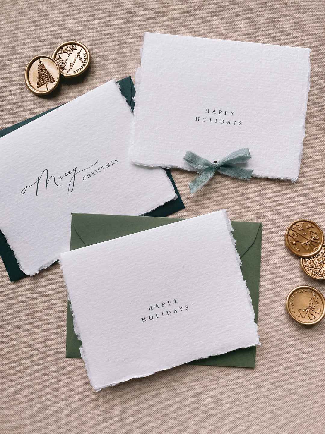 Assorted handmade Christmas cards with two designs of Merry Christmas and Happy Holidays, displayed with olive envelopes and deep green envelopes and one with punched holes with green silk ribbon with Christmas gold wax seals beside the cards
