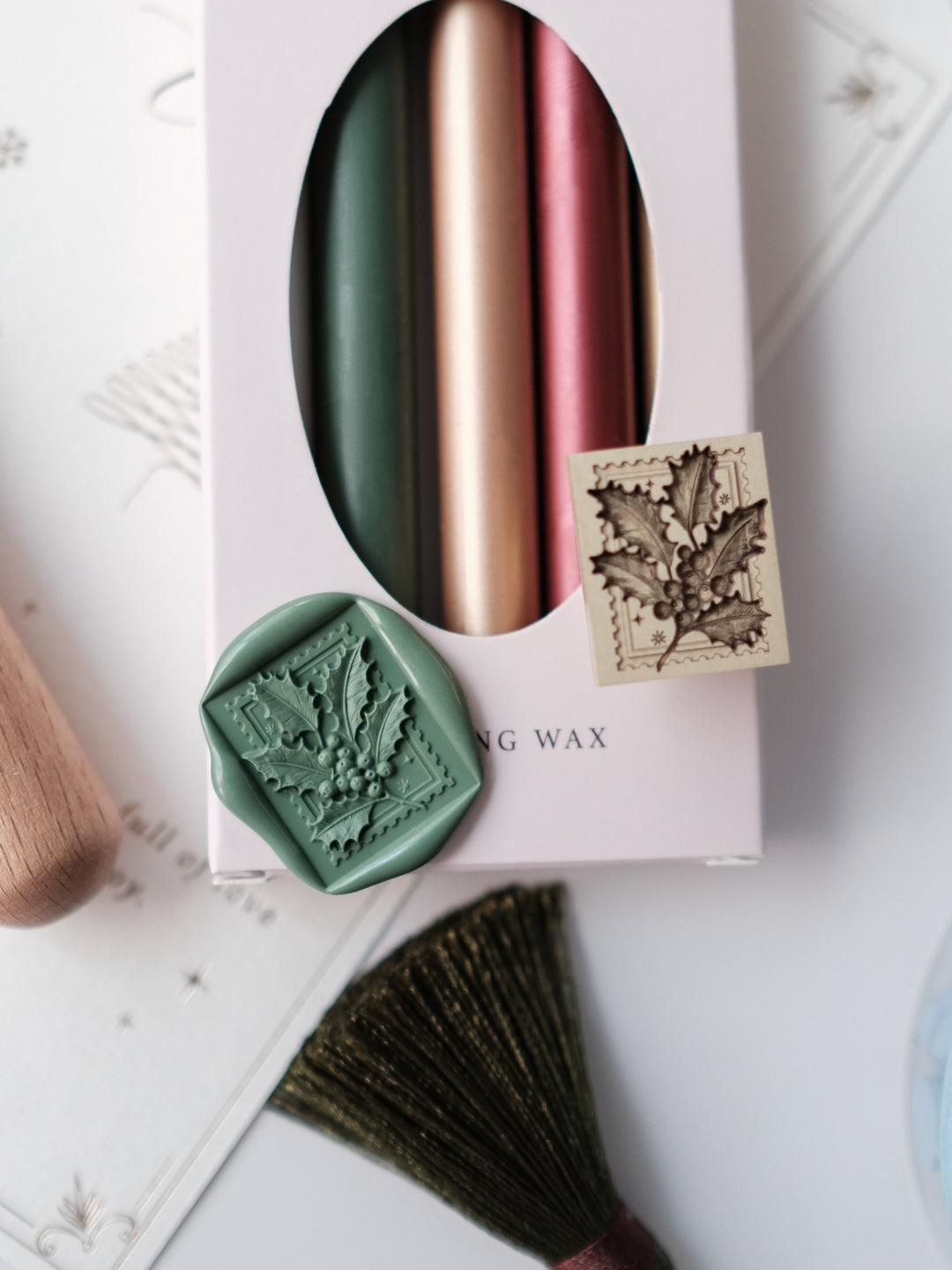 Green Christmas holly leaf wax seal with 3D engraving next to a holly wax stamp head placed on a pack of festive colored sealing wax sticks
