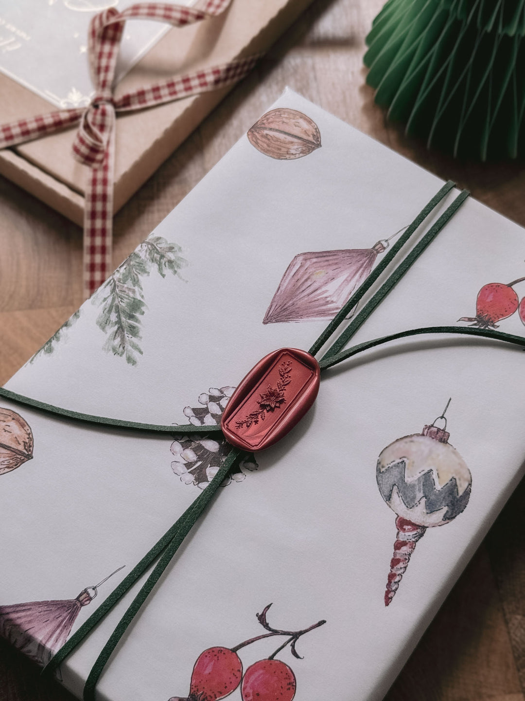 Dark metallic red Christmas poinsettia floral wreath rectangular wax seal on a dark green suede twine tied around a white wrapped gift printed with festive elements 