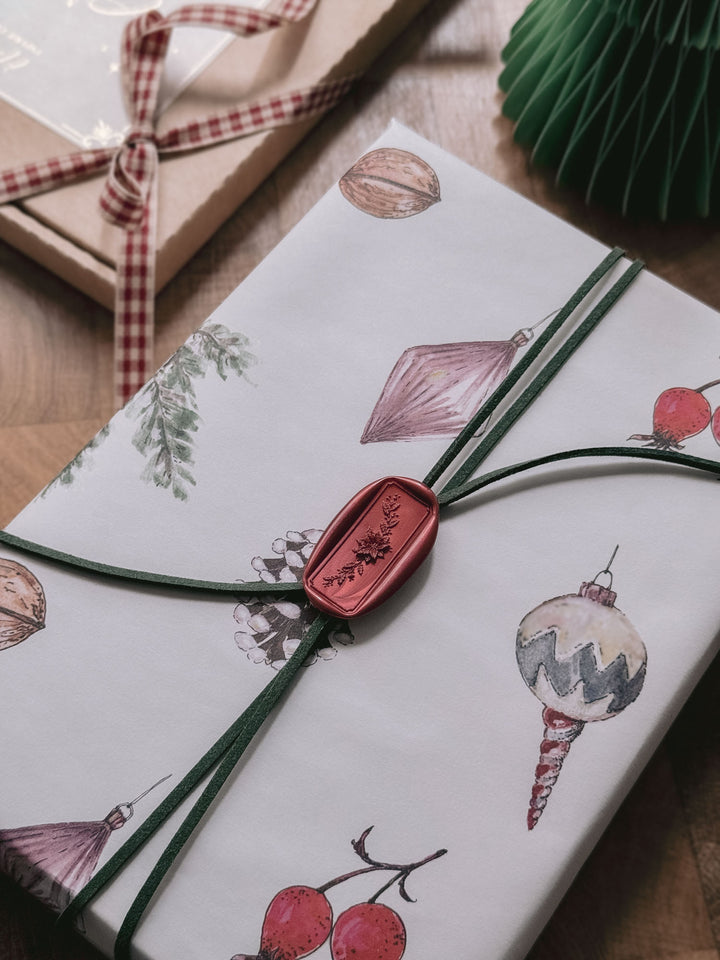 Dark metallic red Christmas poinsettia floral wreath rectangular wax seal on a dark green suede twine tied around a white wrapped gift printed with festive elements 