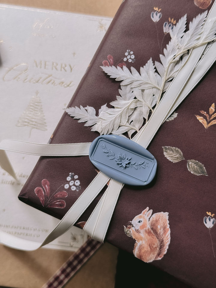 Dusty blue Christmas poinsettia floral garland rectangular wax seal on a white decorative ribbon tied around a brown wrapped gift printed with festive elements 