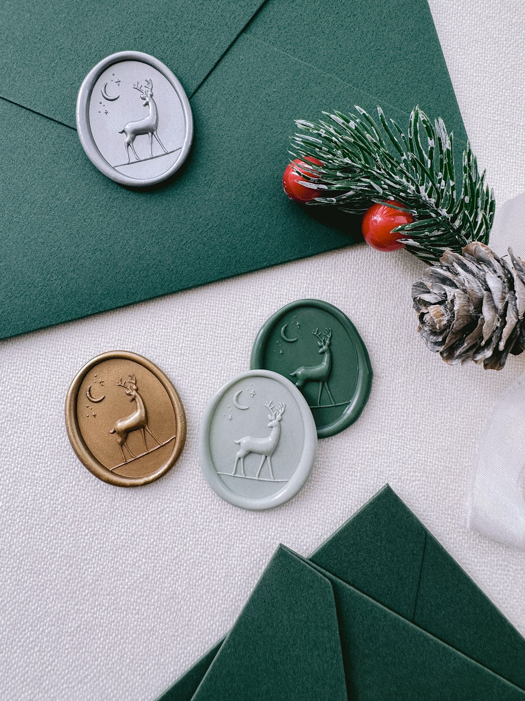 Light sage green, gold, silver and dark green reindeer oval wax seals on a dark green envelope and a light beige background 