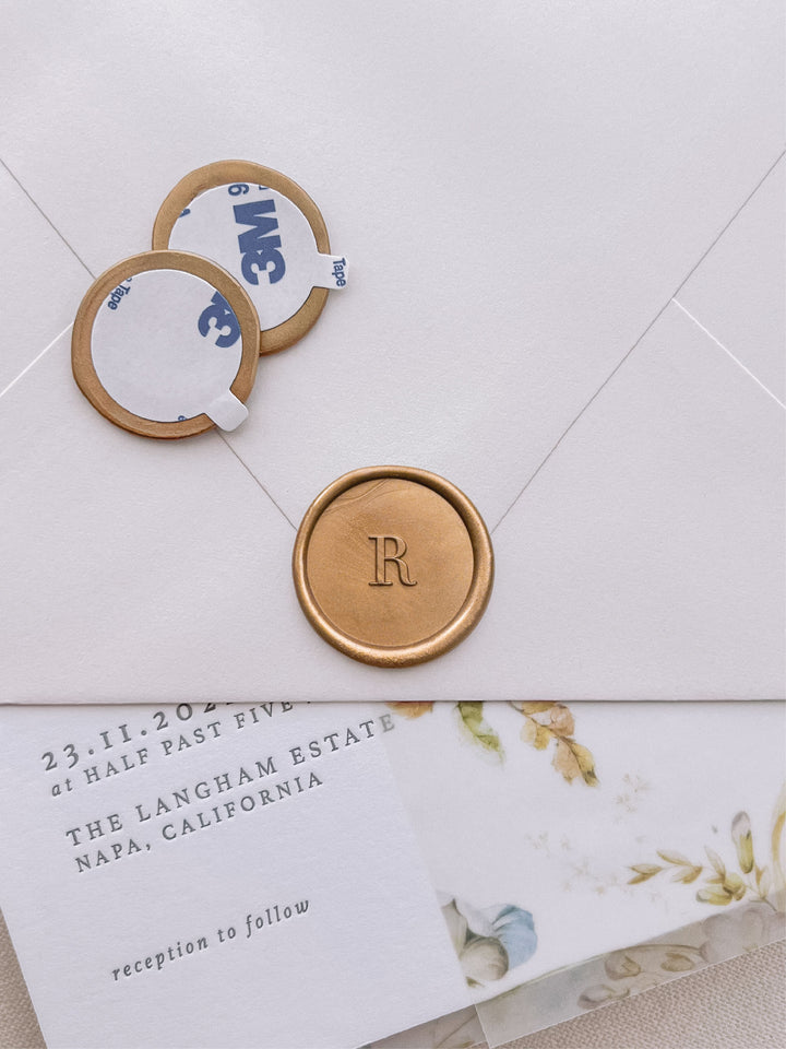 classic initial monogram gold wax seal stickers with self-adhesive backings on a wedding invitation, beige envelope, and floral vellum jacket