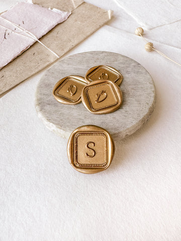Border mini square single initial custom wax seals in gold styled with a small gray stone dish, handmade paper and a dried floral branch