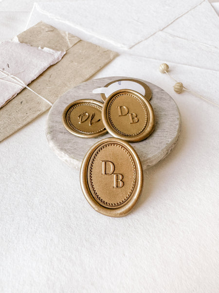 Border monogram oval gold custom wax seals in gold styled with a small gray stone dish, handmade paper and a dried floral branch