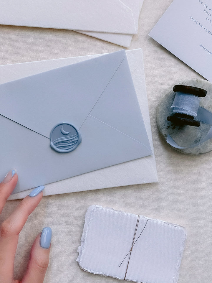 Moon and ocean waves with 3D engraving wax seal in dusty blue on light blue paper envelope styled with handmade paper place cards tied with fine twine and a spool of silk ribbon